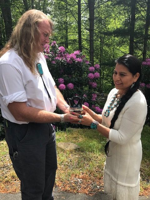 Two people exchanging a small object outdoors near purple flowers and trees