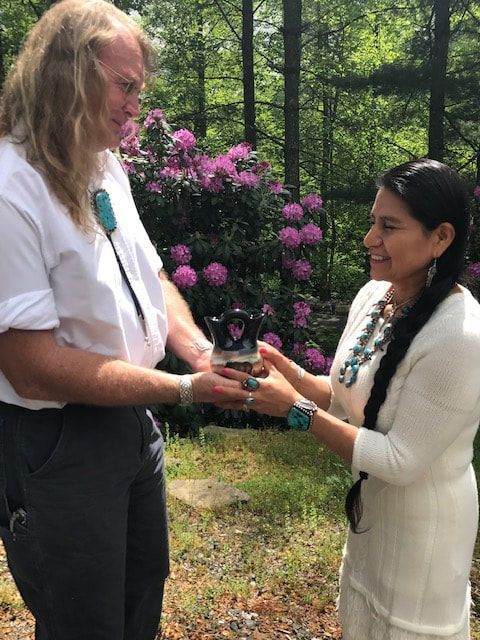 Two people exchanging a small object outdoors near pink flowers and trees, smiling at each other
