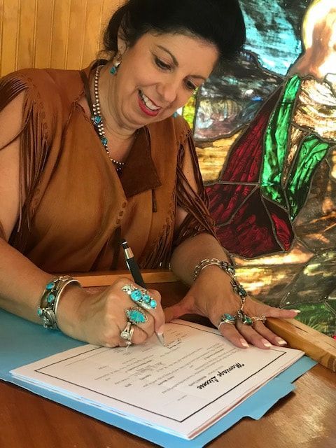 Woman smiling and signing a document at a table, wearing jewelry, with colorful stained glass behind her