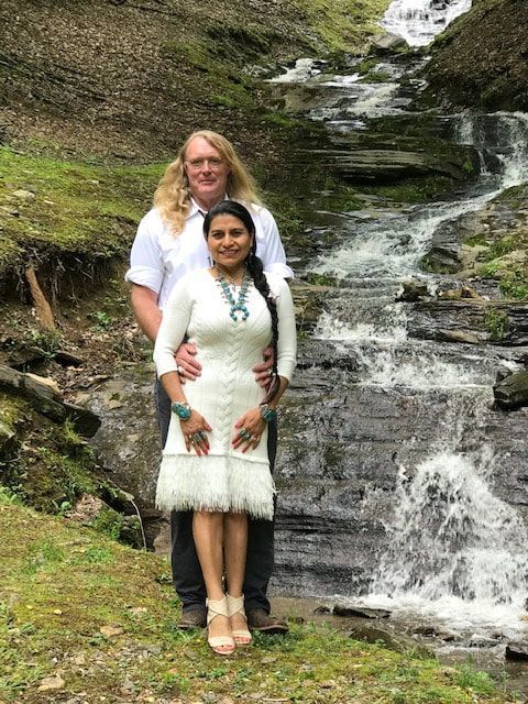 Two people standing by a waterfall on a rocky hillside, one in a white dress and one in a white shirt.