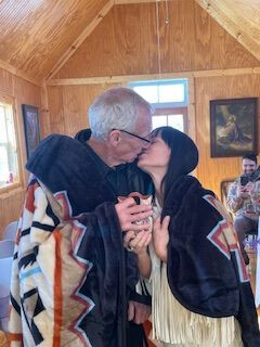 Two people wrapped in a blanket kiss inside a wooden room, with a painting in the background.