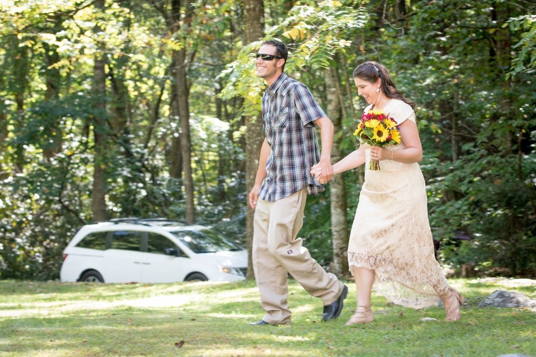 A couple walking hand in hand on a grassy path, with trees and a white car in the background.