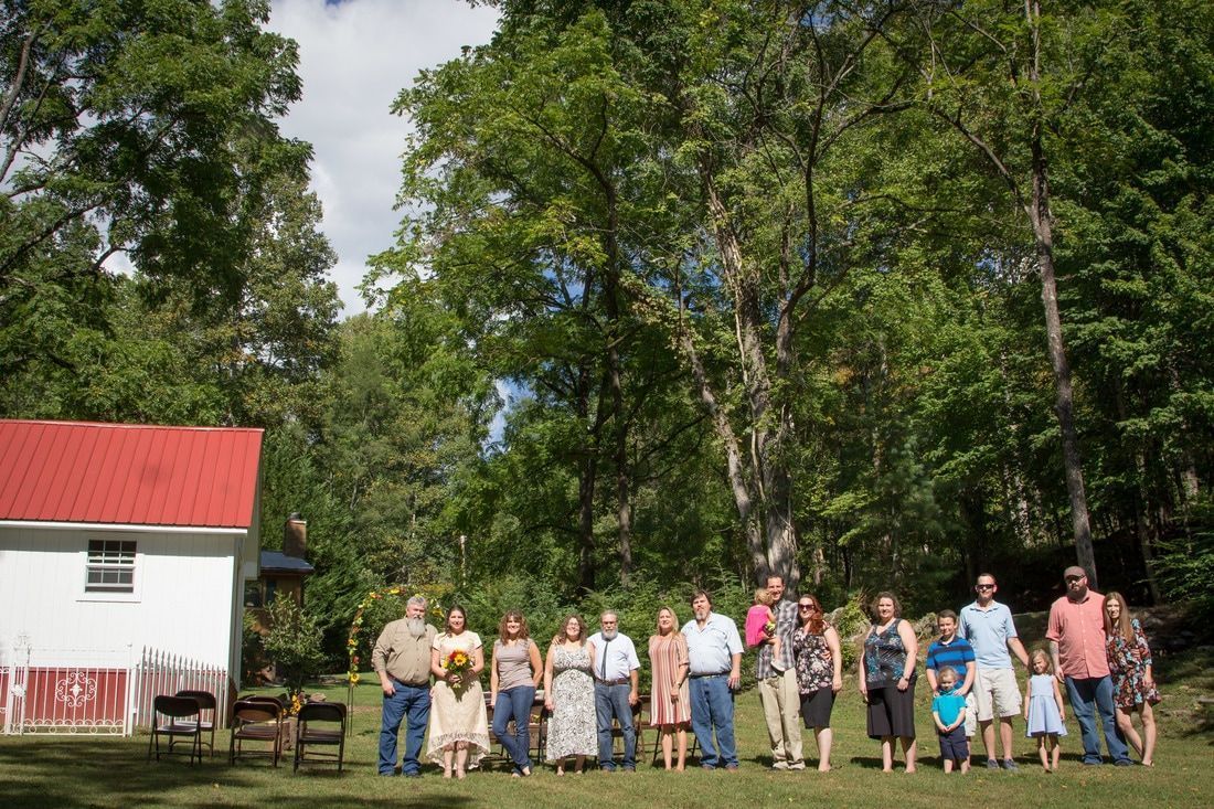 Group of people standing on grass beside a white building with a red roof, surrounded by tall trees.