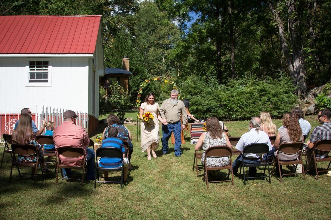 Outdoor gathering with seated audience facing two people standing on grass beside a white building and trees