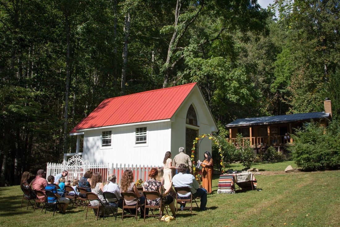 People seated outdoors near a small white building with a red roof in a wooded yard