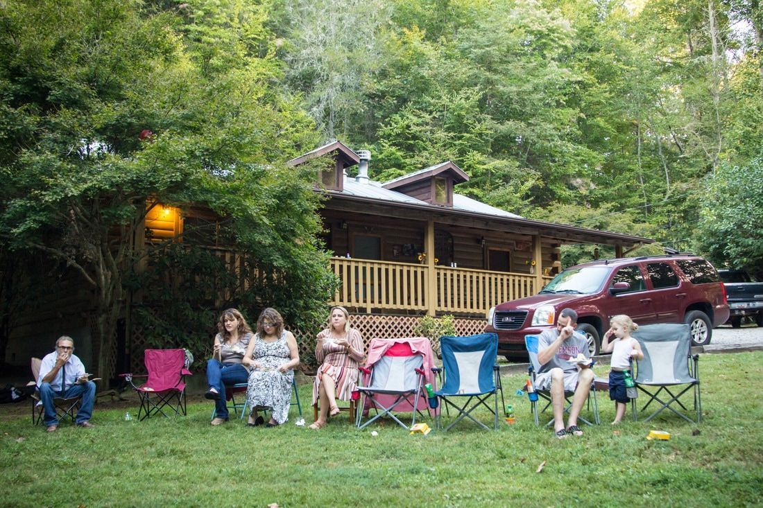 Group seated in camping chairs on a grassy lawn beside a cabin and parked SUV in a wooded area