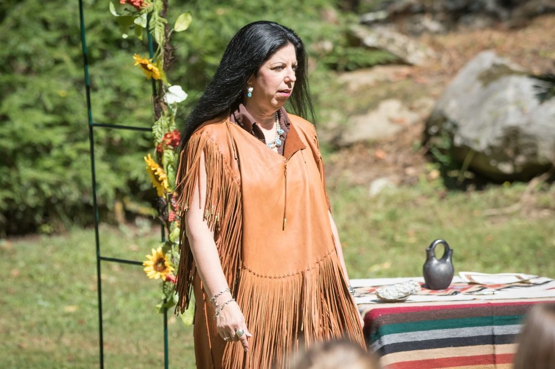 Woman in an orange fringed top stands outdoors beside a striped table with a vase.