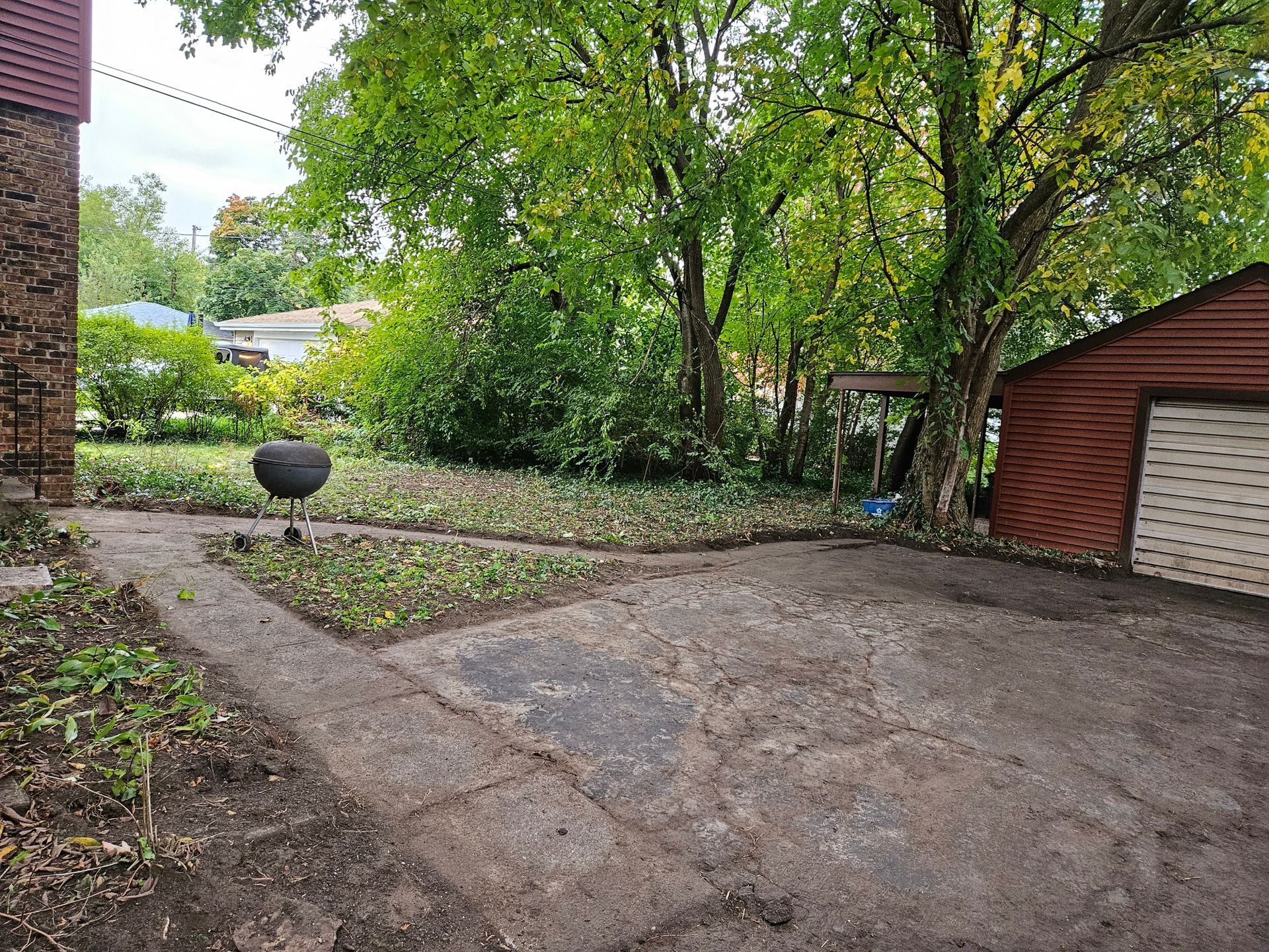 Backyard with bare dirt path, leafy trees, and a red shed beside a garage and garden bed