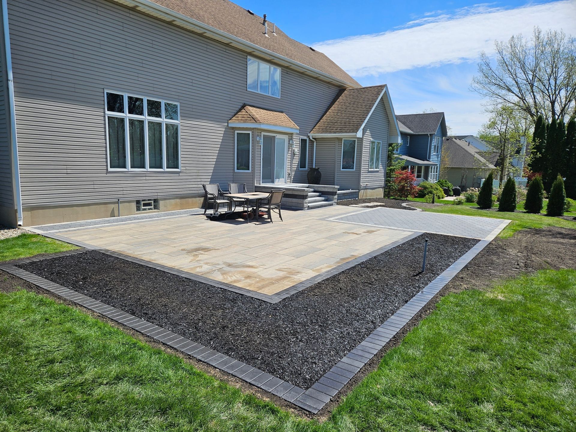 Patio and outdoor seating beside a gray suburban house with a stone border and green lawn