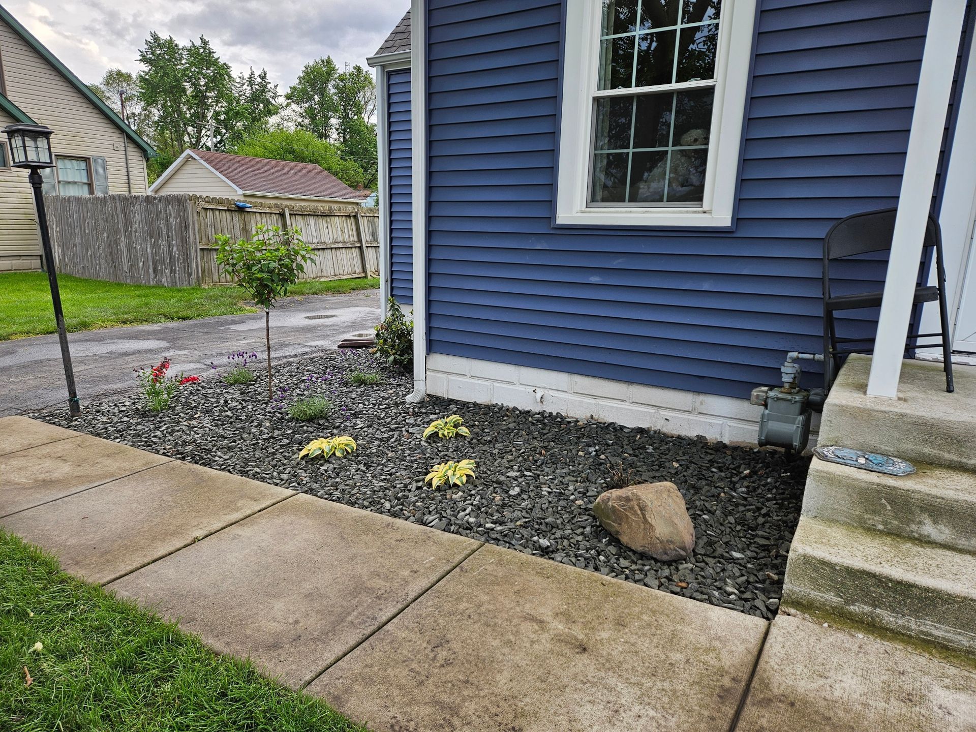 Side of a blue house with a window, concrete walkway, gravel bed, and steps by the entrance.
