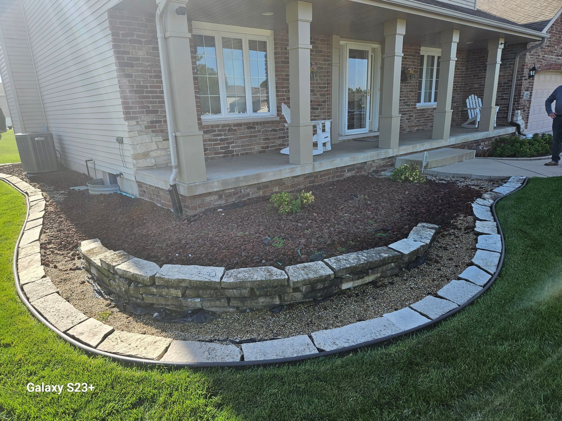 Curved stone garden bed beside a brick house with a walkway and lawn