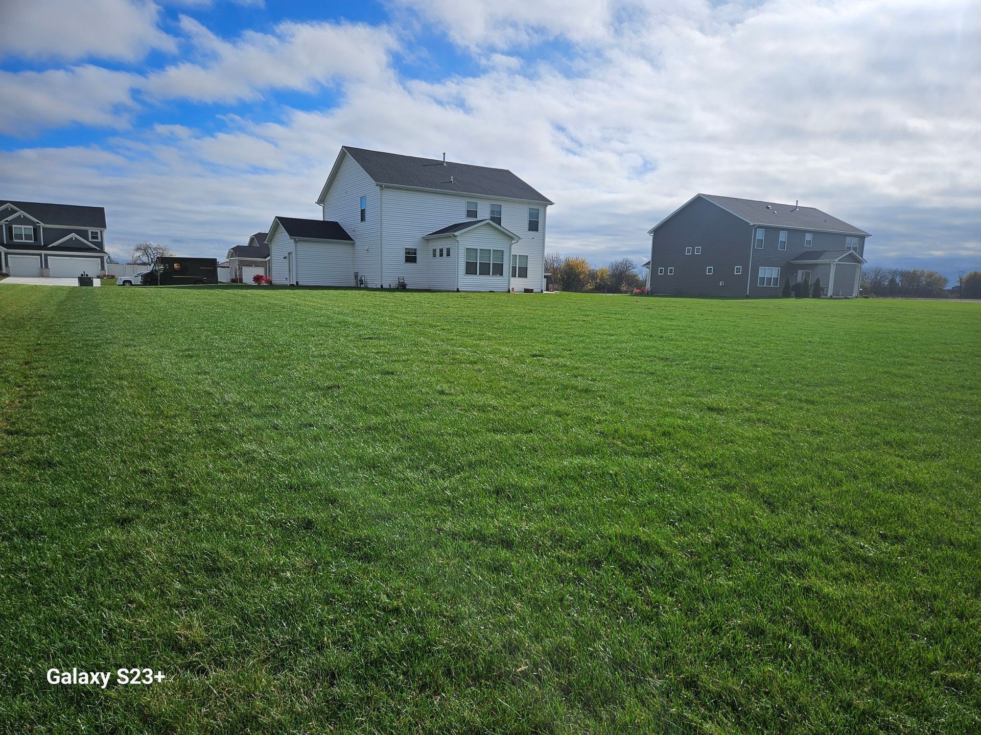 Wide green field with houses under a cloudy blue sky.