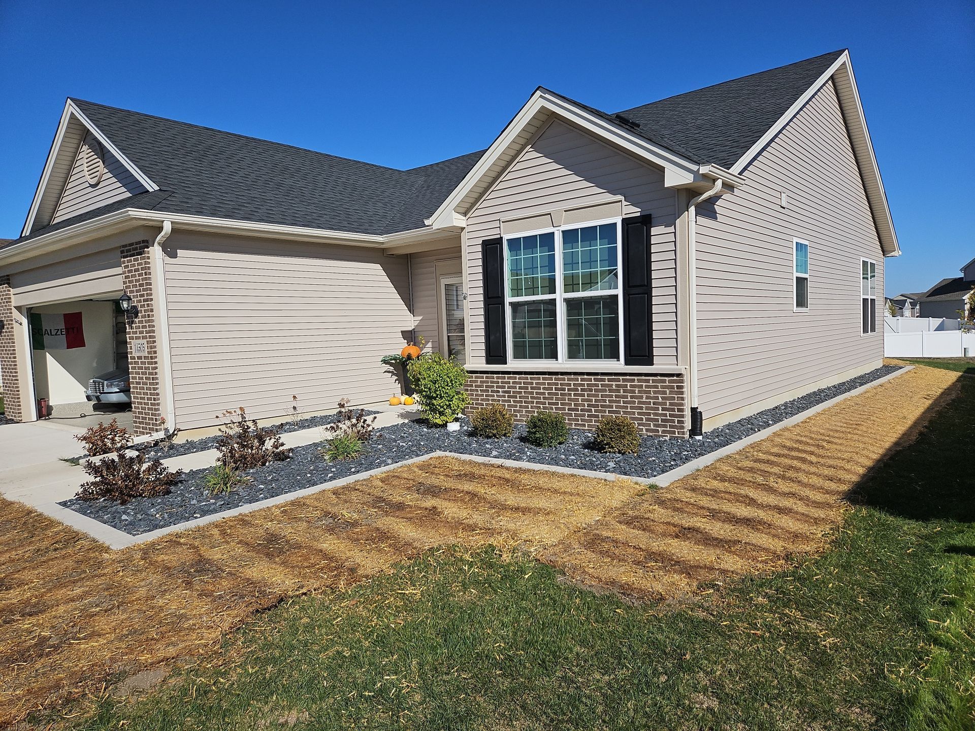 Front yard with a leafless tree, green grass, and a townhouse exterior with a patio and small garden beds.