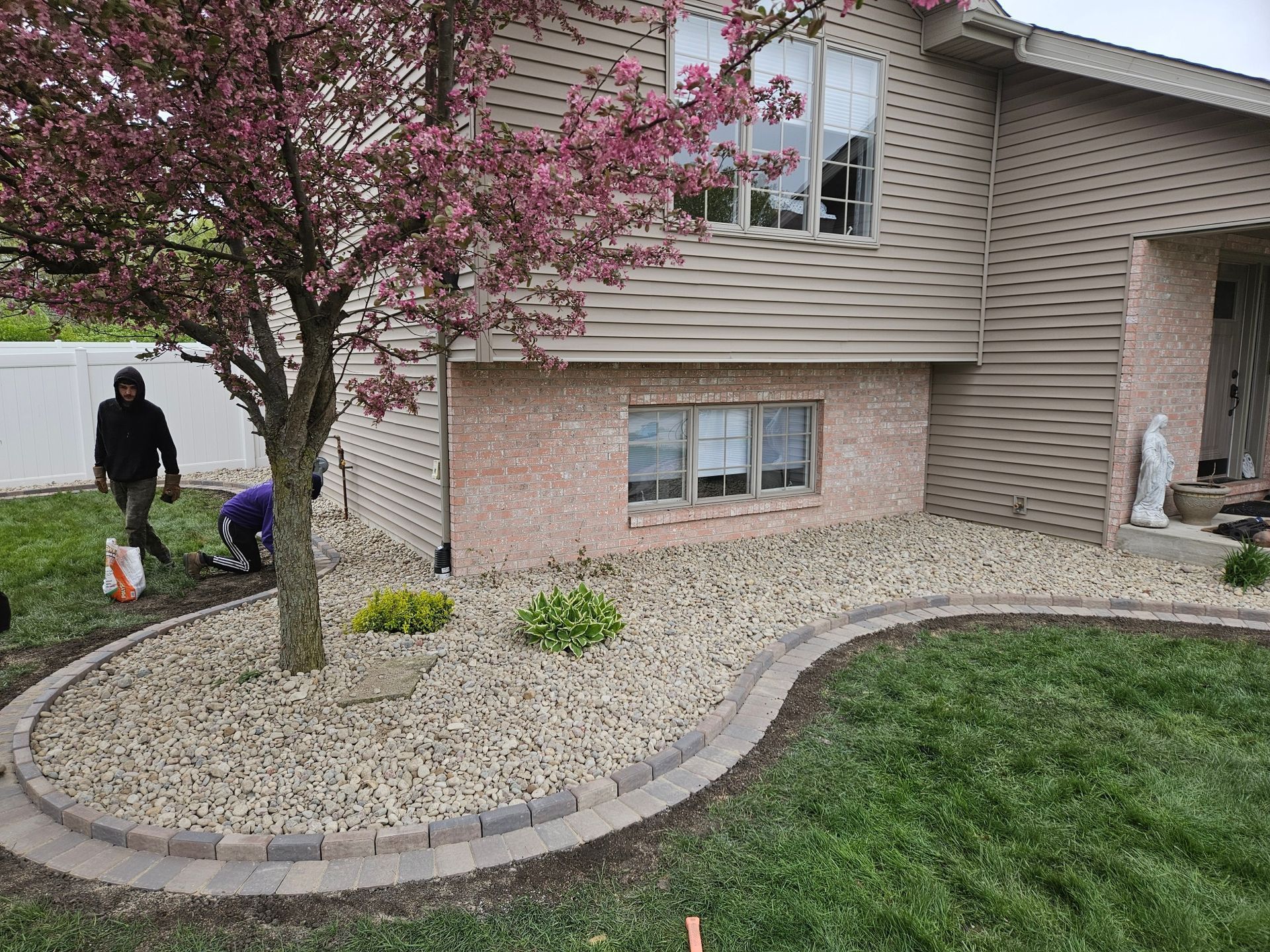 Two workers near a pink flowering tree in a front yard with a curving rock border and brick house.
