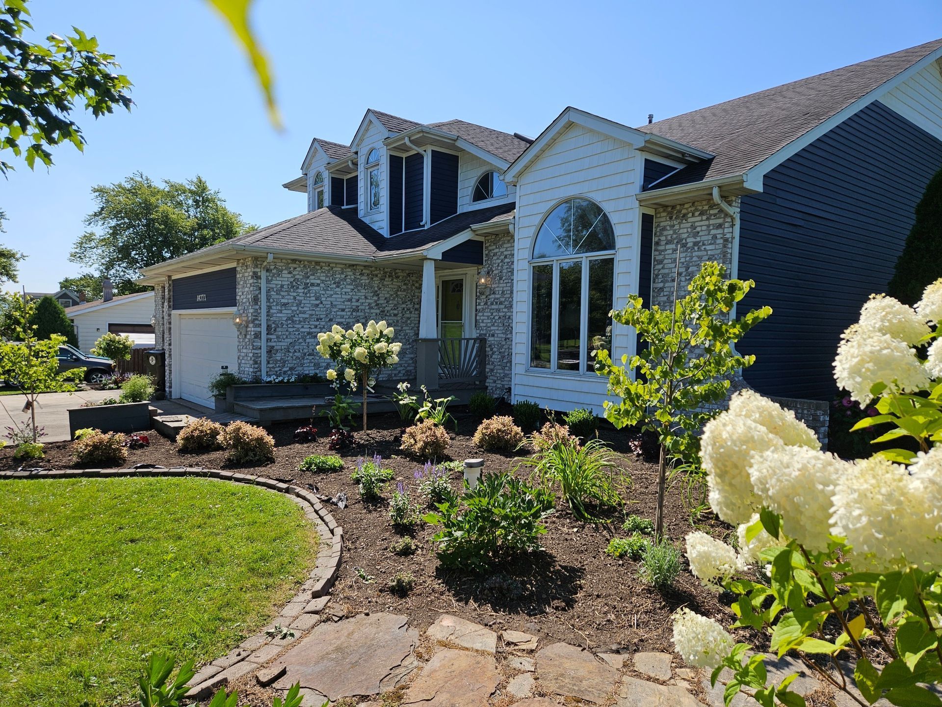 Front yard of a suburban house with white columns, stone facade, and landscaped garden in bright sunlight