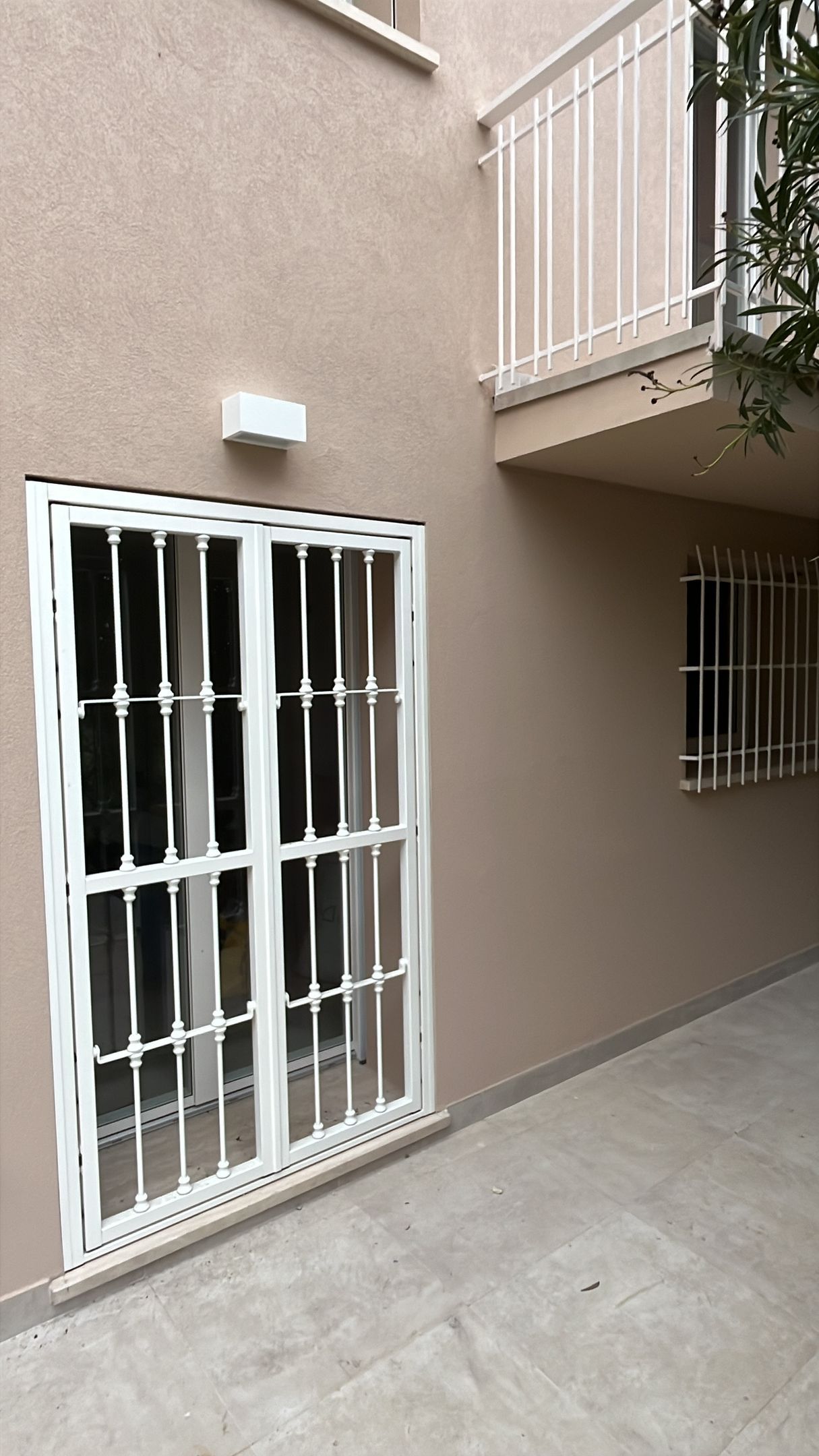 Cortile stretto con muri beige, finestre con inferriate, pavimento in cemento e balcone sovrastante.