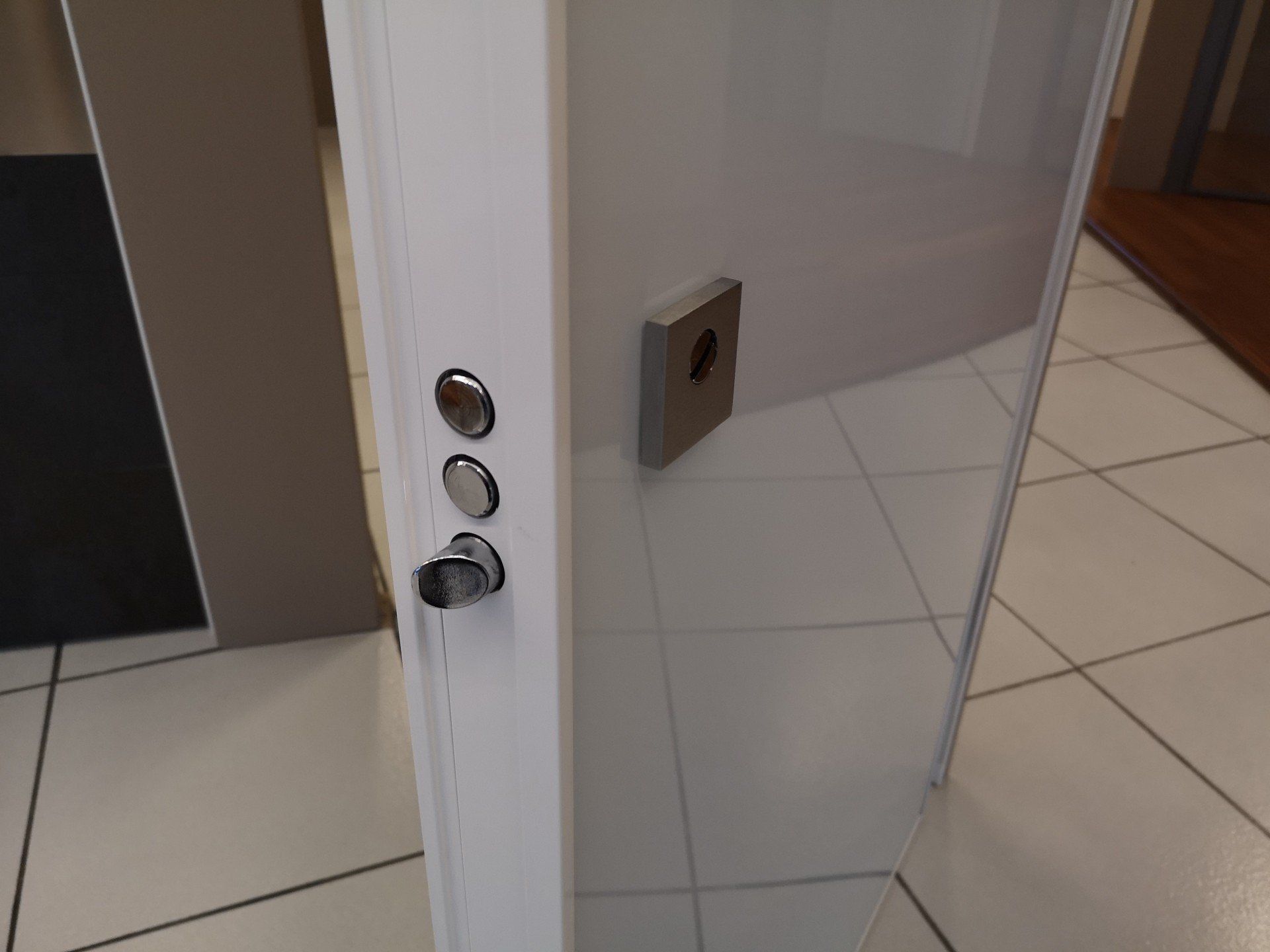 Porta a vetri parzialmente aperta con maniglia e serratura argentate in un corridoio piastrellato.