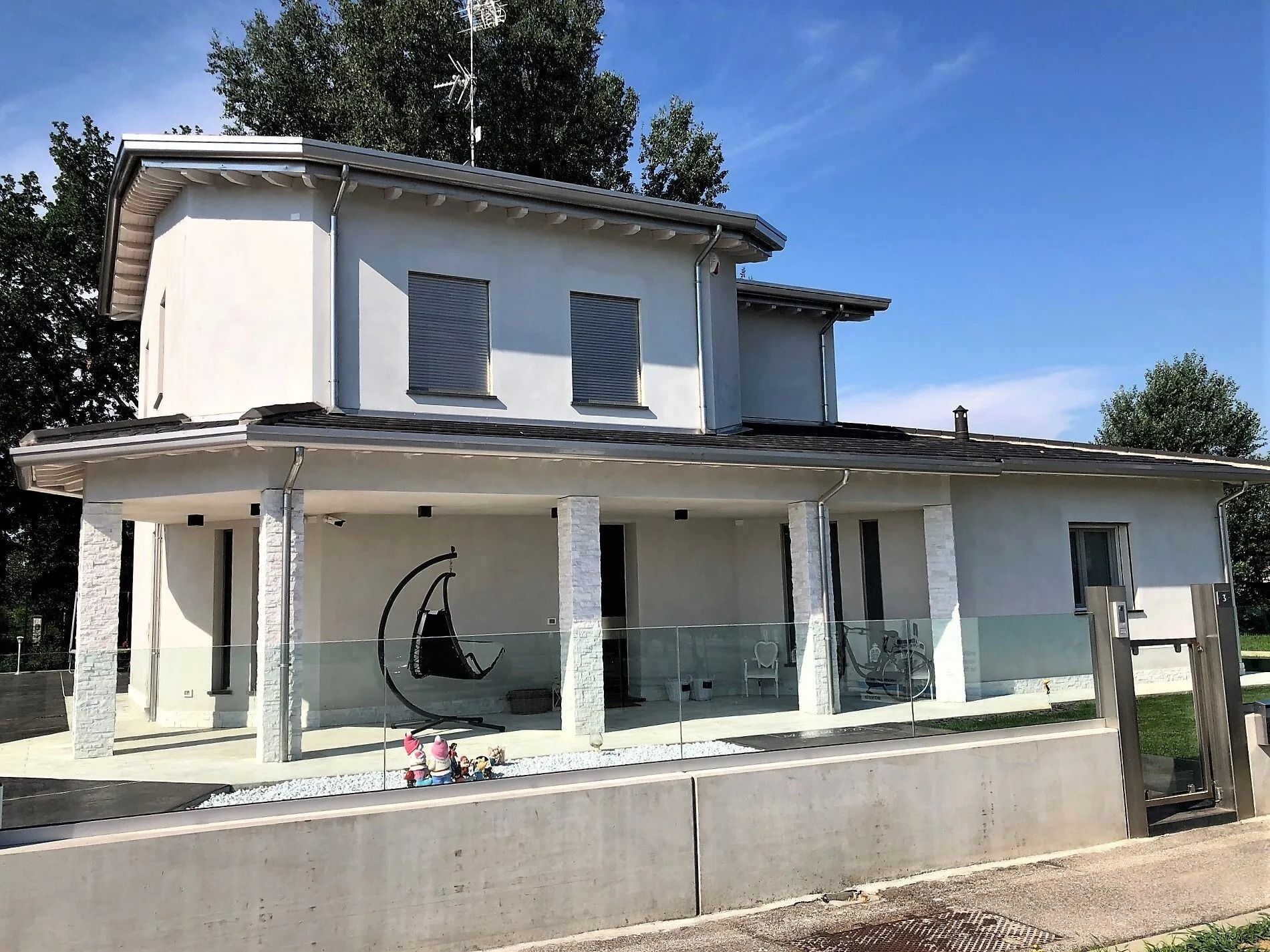 Casa bianca a due piani con portico coperto e ringhiere in vetro sotto un cielo azzurro limpido.
