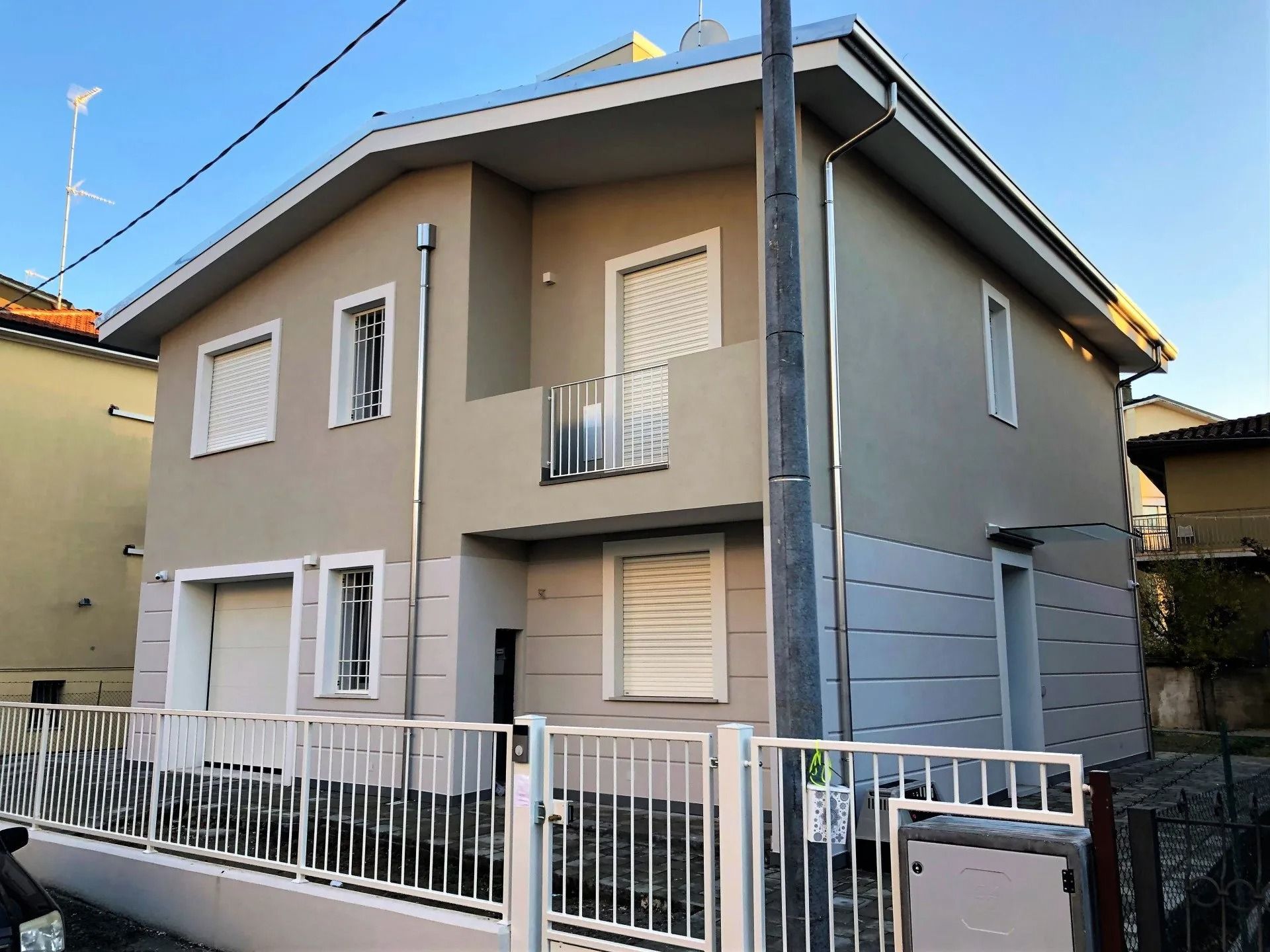 Casa beige a due piani con finiture bianche, balcone e recinzione anteriore in una giornata di sole.