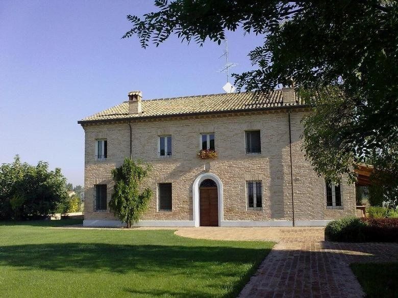 Casa in pietra con porta ad arco in legno e finestre con persiane, immersa in un giardino soleggiato.