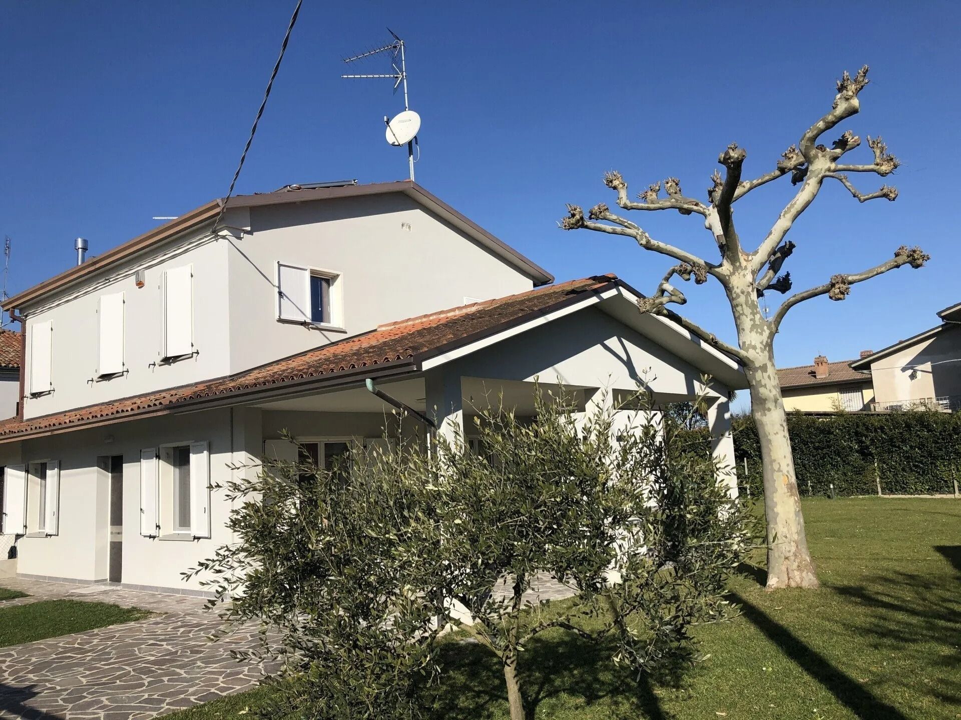 Casa bianca a due piani con tetto rosso e giardino, accanto a un albero potato sotto un cielo limpido