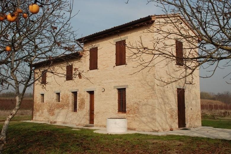 Vecchia casa colonica beige con persiane rosse, immersa in un prato rurale e incorniciata da alberi spogli.