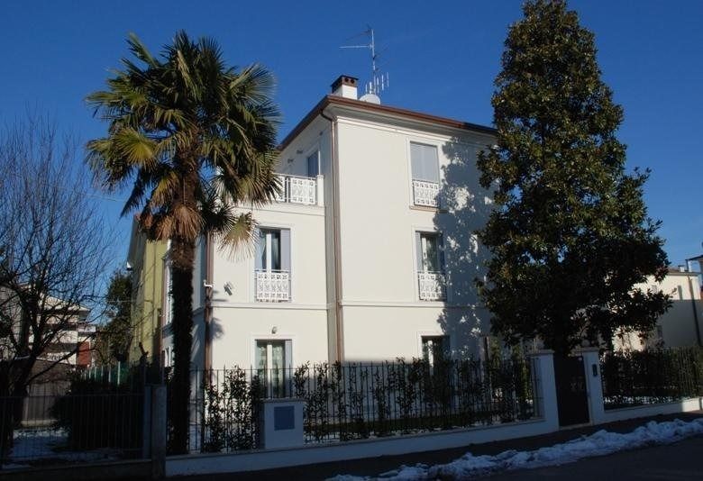 Casa color crema con tetto rosso, palma e alti alberi sempreverdi dietro una recinzione in ferro nero.