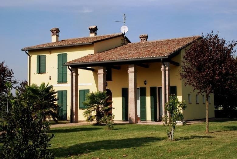 Casa di campagna gialla con persiane verdi, tetto di tegole e veranda ombreggiata circondata da erba e alberi.