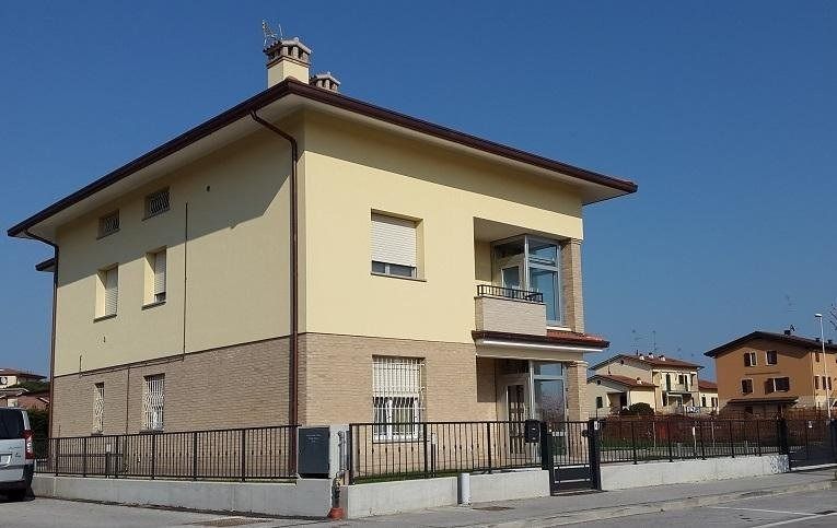 Casa a due piani color crema con balcone e cortile recintato, situata in una strada soleggiata.
