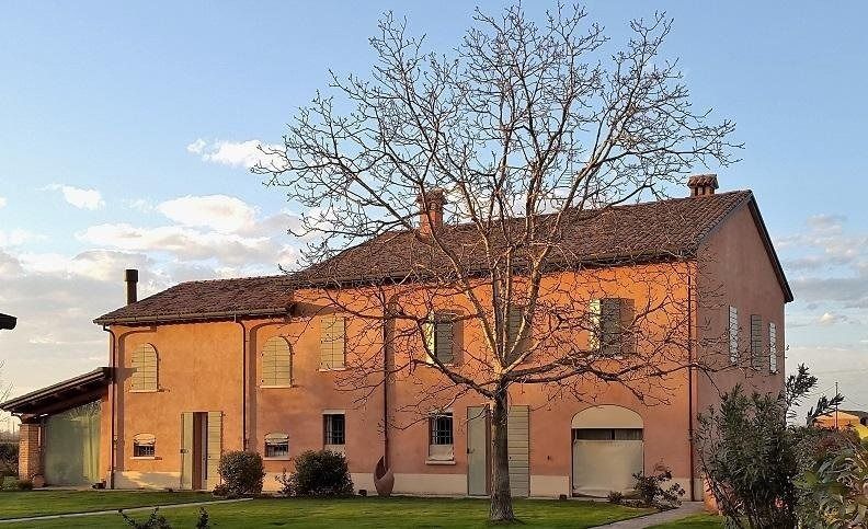Casa a due piani color beige con tetto di tegole rosse e albero spoglio di fronte, sotto un cielo azzurro.