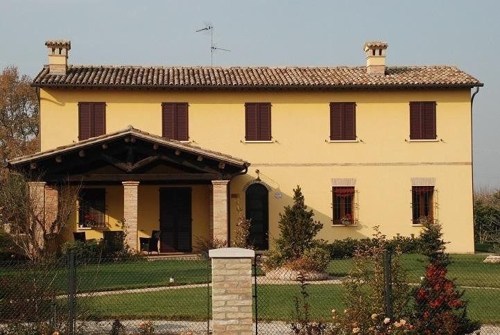 Casa gialla a due piani con persiane scure, portico coperto e giardino anteriore dietro una recinzione.