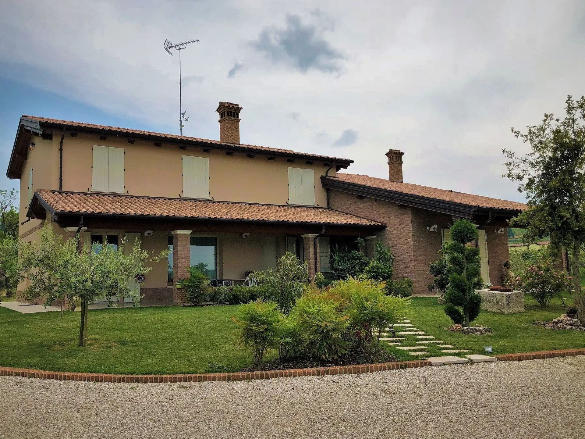 Casa rustica a due piani con tetto di tegole, prato verde e vialetto di ghiaia sotto un cielo nuvoloso