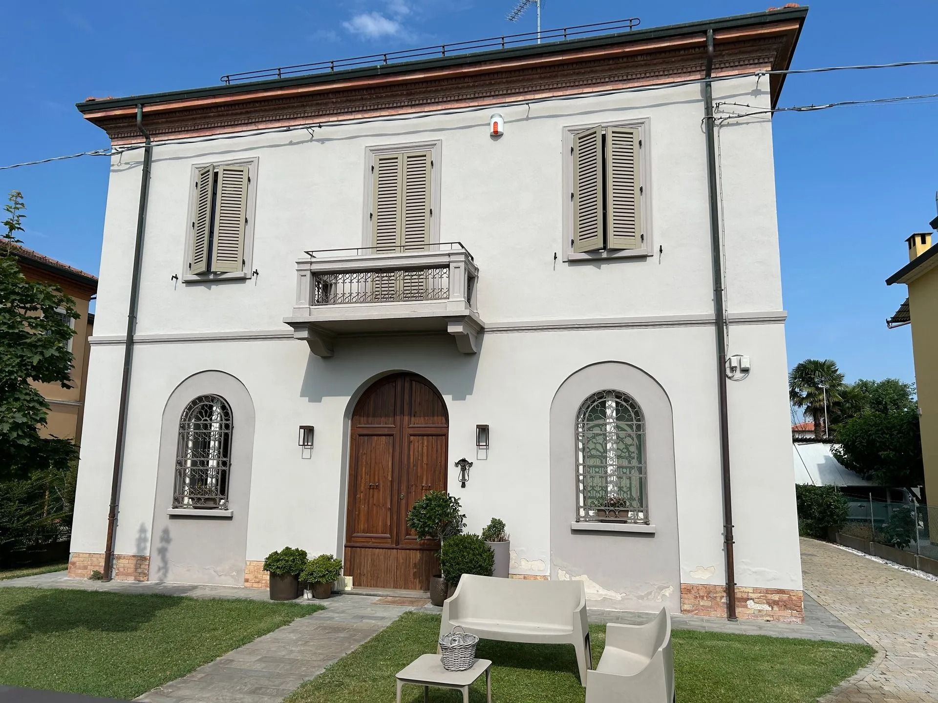 Casa bianca a due piani con finestre ad arco, porta d'ingresso in legno, balcone e prato verde sotto un cielo azzurro.