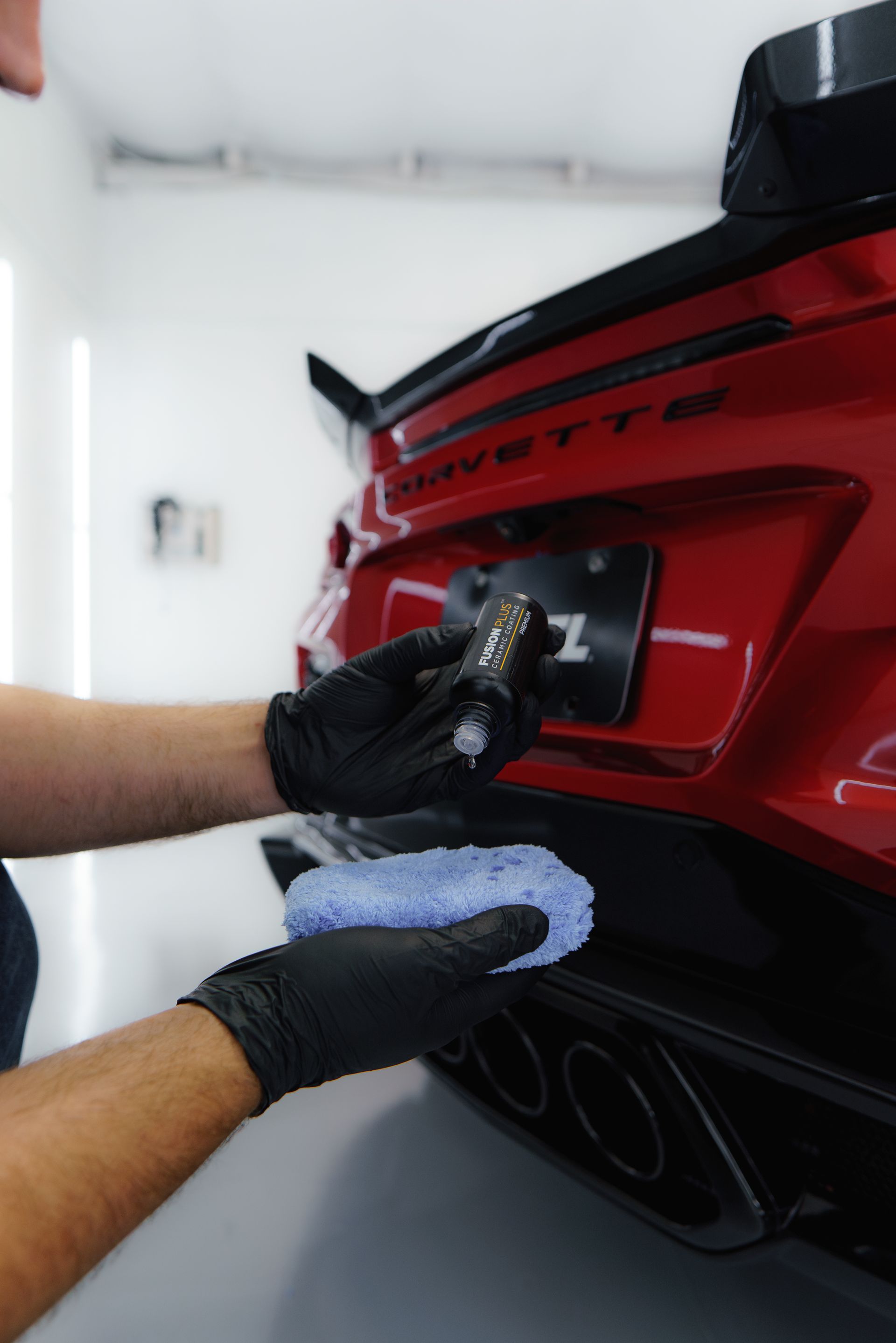 Person in black gloves applying detailing product to a red Corvette's bumper with a blue applicator in a garage.