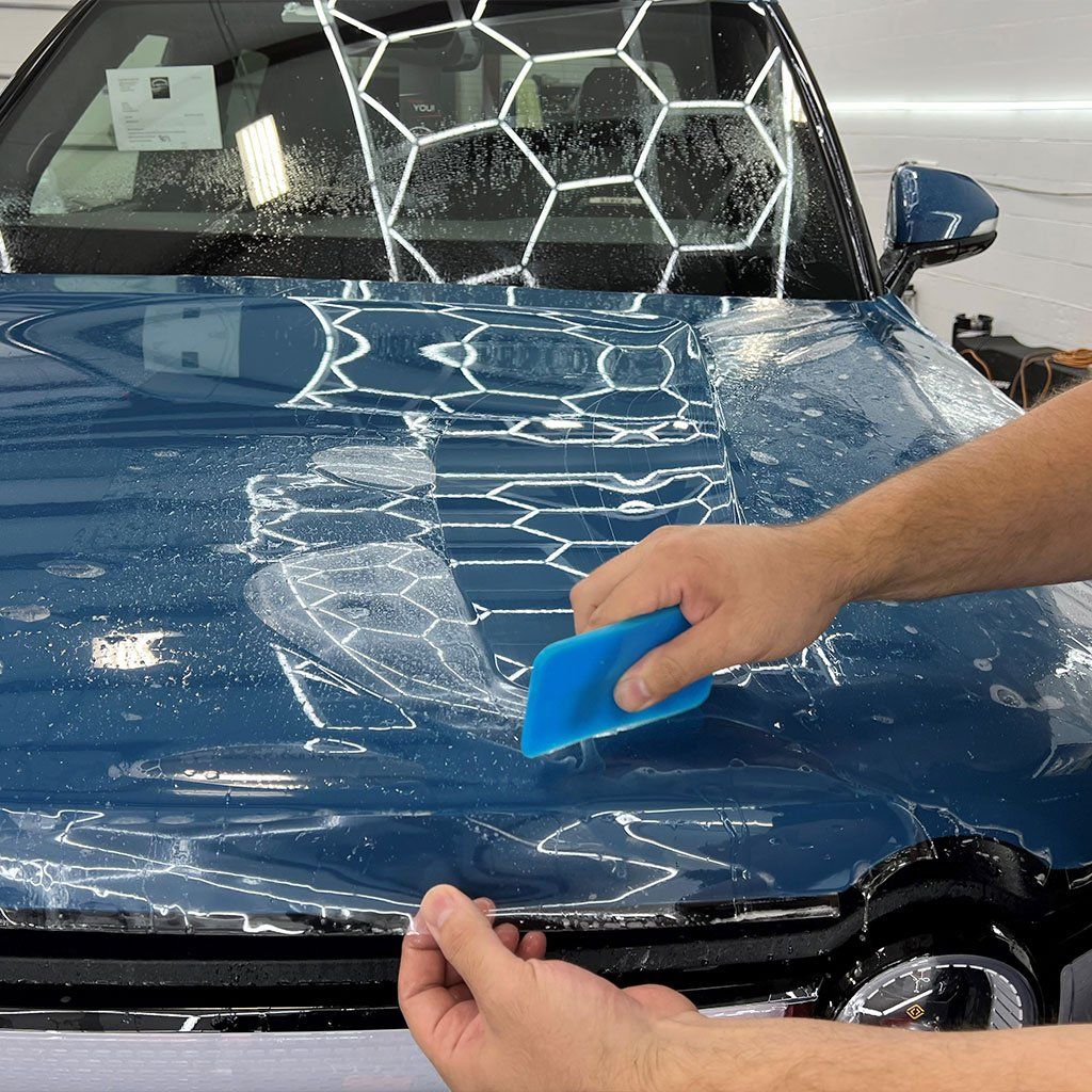 Person applies protective film to a blue car hood using a blue squeegee in a well-lit garage.