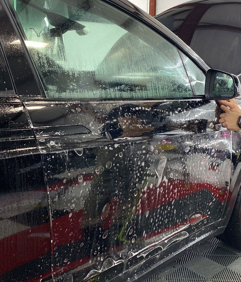 A person applying a protective film to a black car's front door in a well-lit workshop.
