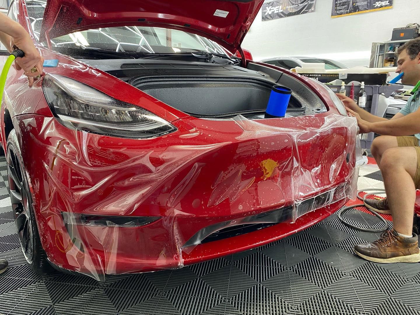 People applying clear protective film to the front of a red Tesla in a shop.