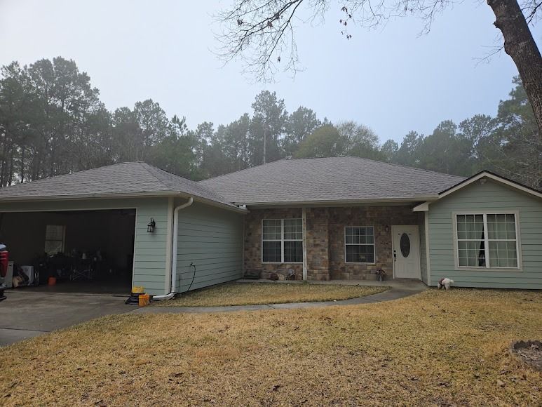 Single-story house with light green siding, brick center, and an open garage on a wooded lot