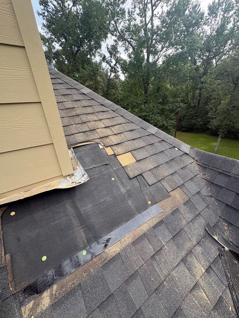 Damaged shingled roof with missing and exposed patches beside a tan house wall