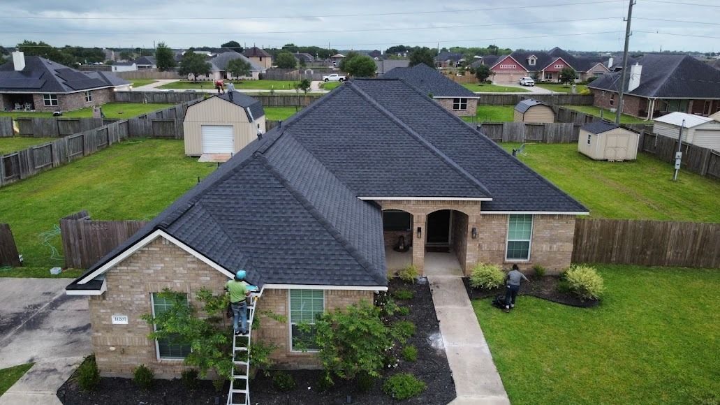 Aerial view of a single-story house with a dark roof, front yard, and fenced neighborhood around it.