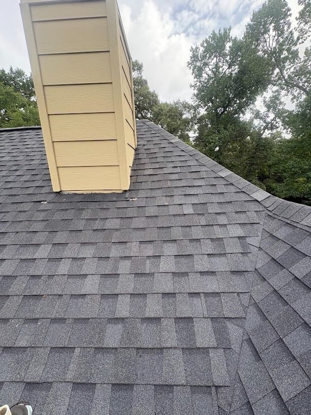Gray shingle roof with a tan chimney and trees in the background