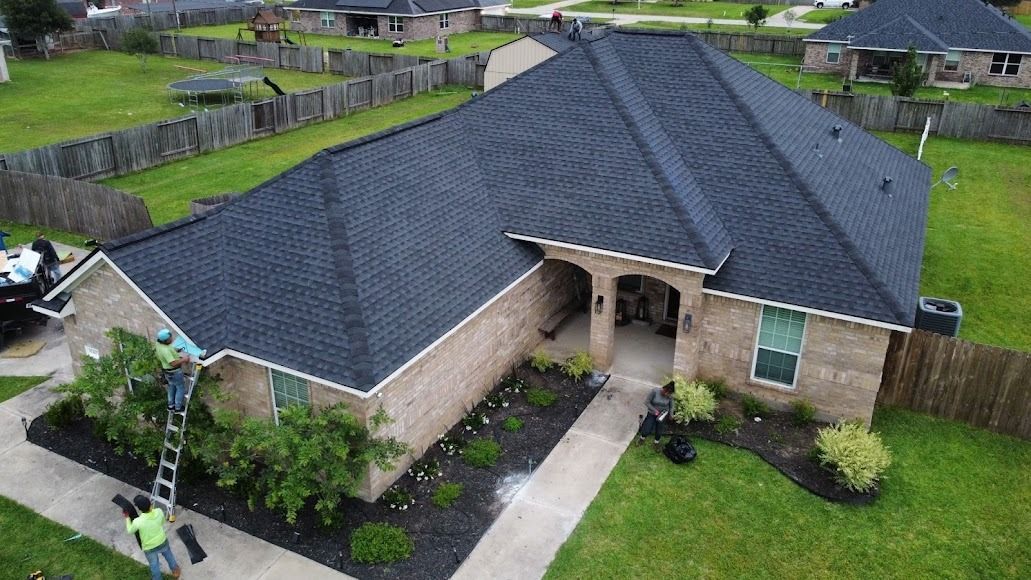 Aerial view of a single-story brick house with a dark gray roof and landscaped front yard in a suburban neighborhood