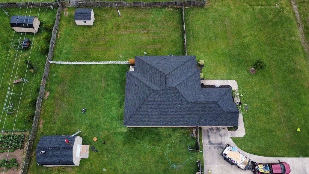 Aerial view of a dark-roofed house with a large green yard, fence, and parked vehicles nearby.