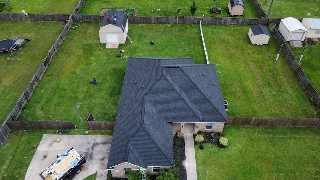 Aerial view of a house with a dark roof, green lawn, and fenced yards in a suburban neighborhood.