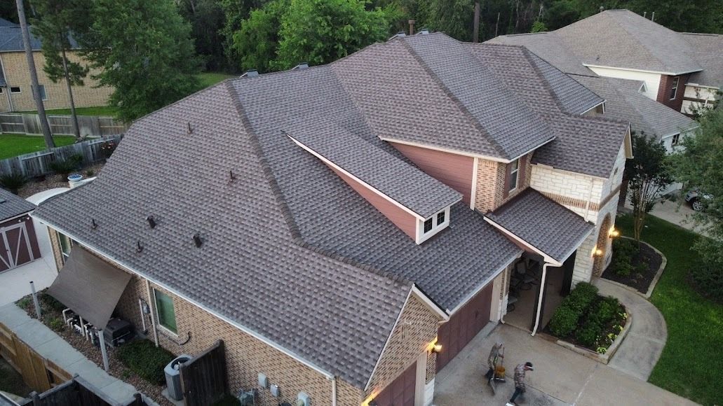 Aerial view of a large suburban house with gray shingled roof and brick exterior, surrounded by trees.