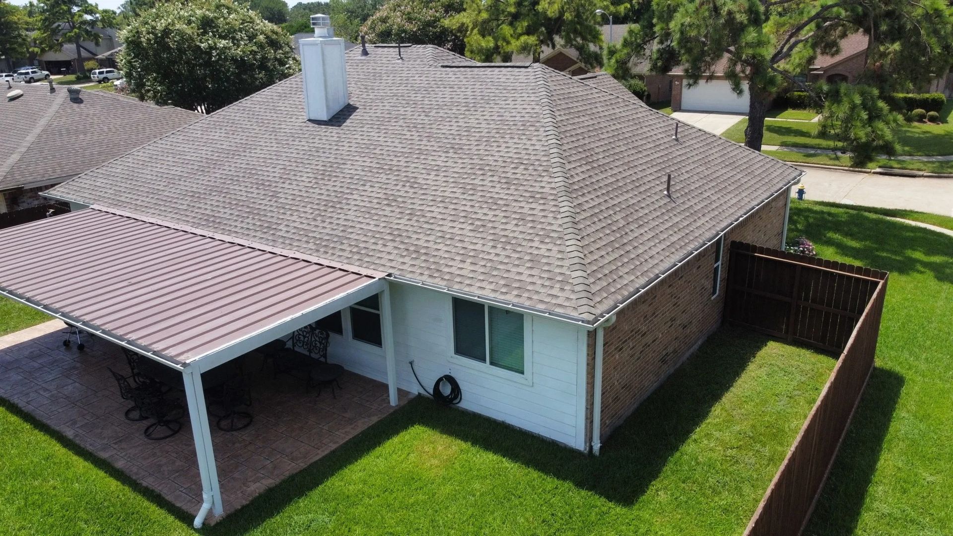 Aerial view of a single-story house with a gray roof, covered patio, and fenced green yard.