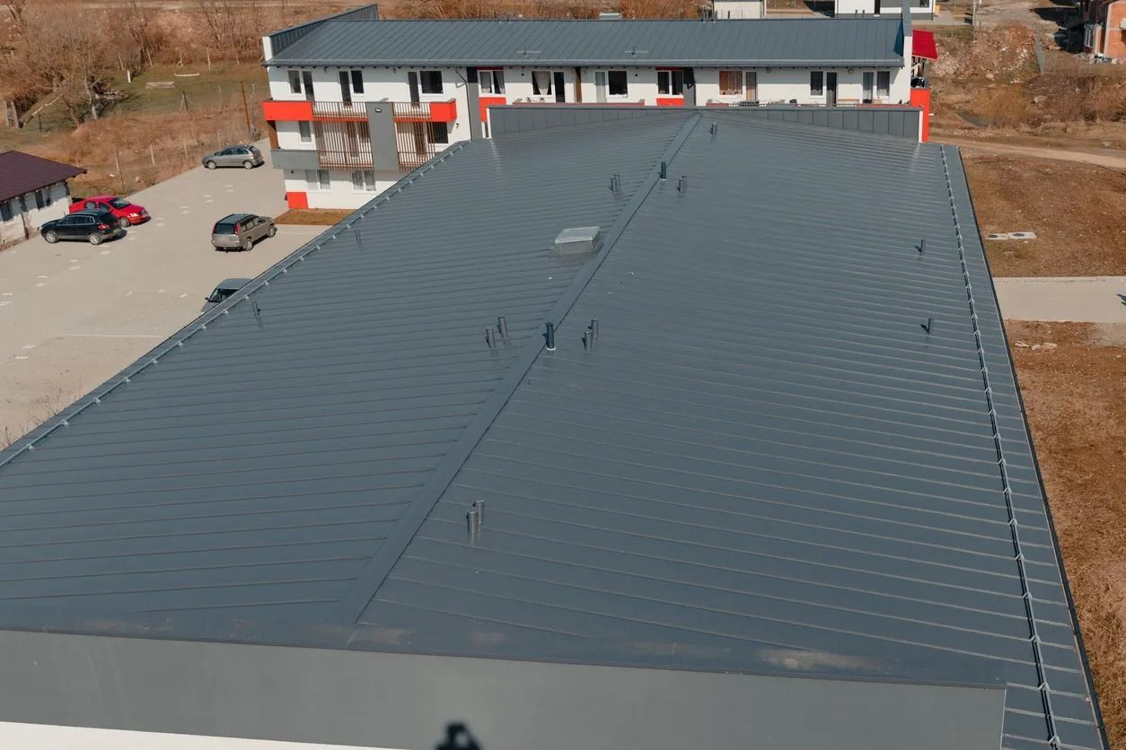 Large gray warehouse roof with skylights, seen from above beside a building and parking area