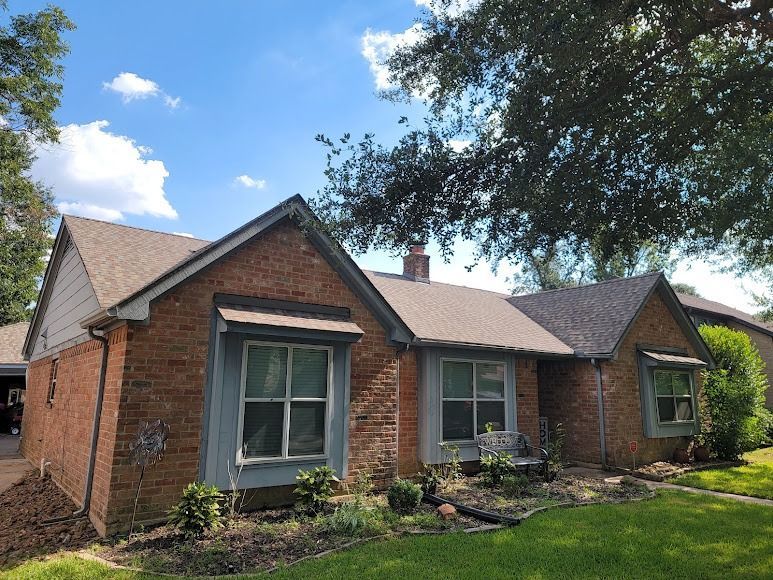 Brick single-story house with gray roof and blue-trimmed windows, front garden, and trees overhead