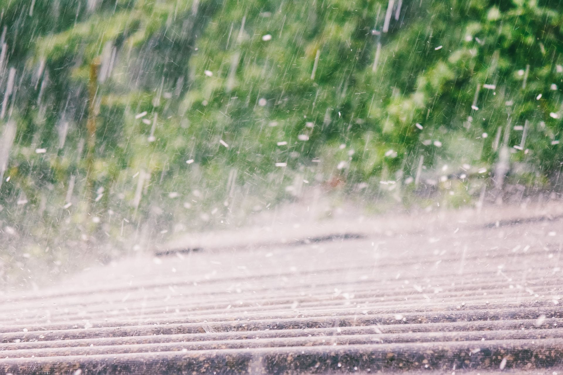 Heavy rain with hail falling onto a roof with blurred green foliage in the background.