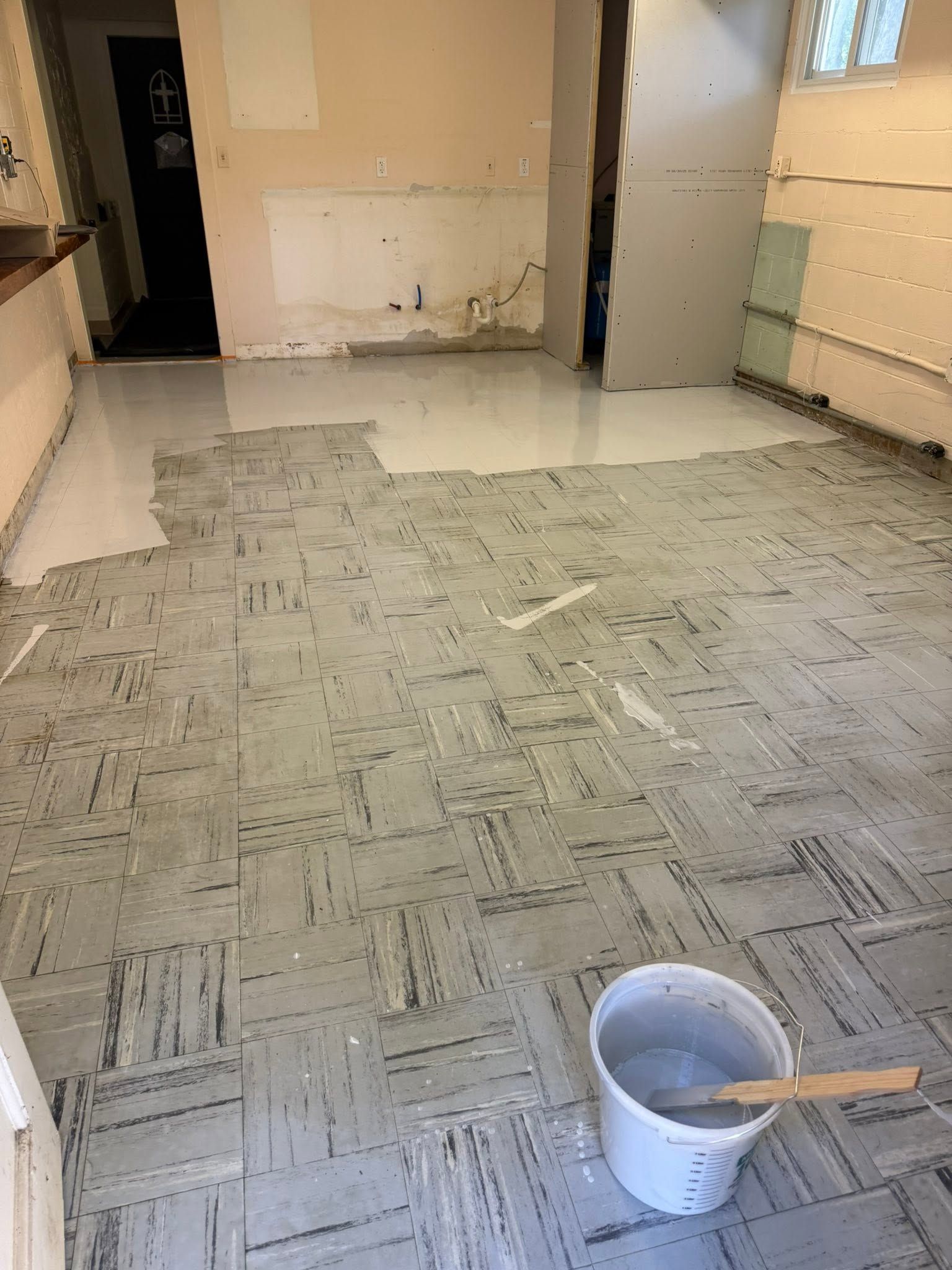 Empty room with new gray tile flooring and a white bucket with a paintbrush in the foreground.